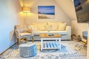 Ein gemütliches Wohnzimmer mit Sofa, Sessel, Hocker und Couchtisch, mit Blick auf den Strand und einer Lampe.