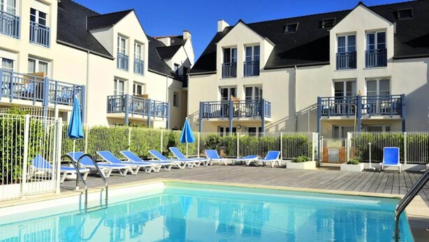 Una piscina con sillones de descanso y sombrillas, rodeada de edificios de apartamentos con balcones.