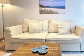 Ein beiges Sofa mit zwei Kissen, ein Couchtisch aus Holz mit einem blauen und weißen Dekorationsgegenstand und ein Gemälde mit Strandblick an der Wand.