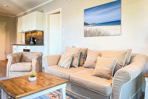 Ein Wohnzimmer mit einem beigen Sofa, einem Couchtisch mit einer Pflanze und einer Küche im Hintergrund.