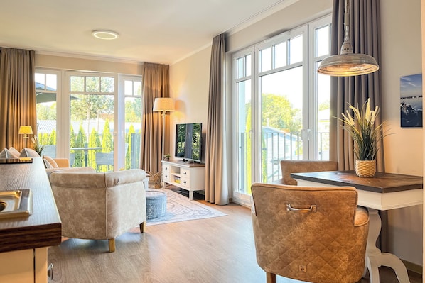 Ein modernes Wohnzimmer mit einer Couch, einem Sessel und einem Esstisch. Es gibt einen Fernseher, eine Lampe und eine Pflanze.