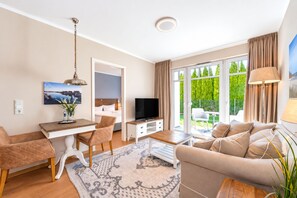 Un salon moderne avec un canapé, une table à manger et un téléviseur. Une grande fenêtre, ornée de rideaux, offre une vue sur la verdure environnante.