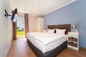 Ein modernes Hotelzimmer mit einem großen Bett, einem flachbildfernseher an der Wand, einem Nachttisch mit Lampe und Blick ins Freie durch eine Schiebetür.