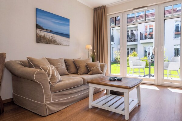 A living room with a sofa, a coffee table, a painting, and a view of a balcony with outdoor furniture. - Baltischer Hof apartment 16 (Ostseebad Boltenhagen)