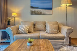 Un soggiorno con un divano beige, un tavolino da caffè, una lampada da terra e un quadro appeso raffigurante una spiaggia.