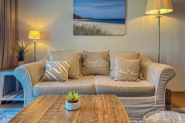 Ein Wohnzimmer mit einem beigefarbenen Sofa, einem Couchtisch, einer Stehlampe und einem an der Wand hängenden Bild von einem Strand.