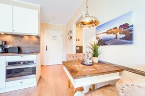 Une cuisine moderne dotée d’appareils électroménagers intégrés, un coin repas avec une table et des chaises en bois, et un grand tableau représentant un paysage marin accroché au mur.