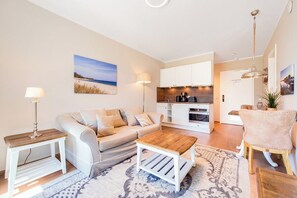 A modern living room with a sofa, coffee table, and lamp. There is a kitchen area with cabinets and a dining area with a chair. - Baltischer Hof 26 Apartment (Ostseebad Boltenhagen)