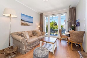 A living room with a sofa, a coffee table, a lamp, a chair, and a view through a glass door. - Baltischer Hof apartment 37 (Ostseebad Boltenhagen)