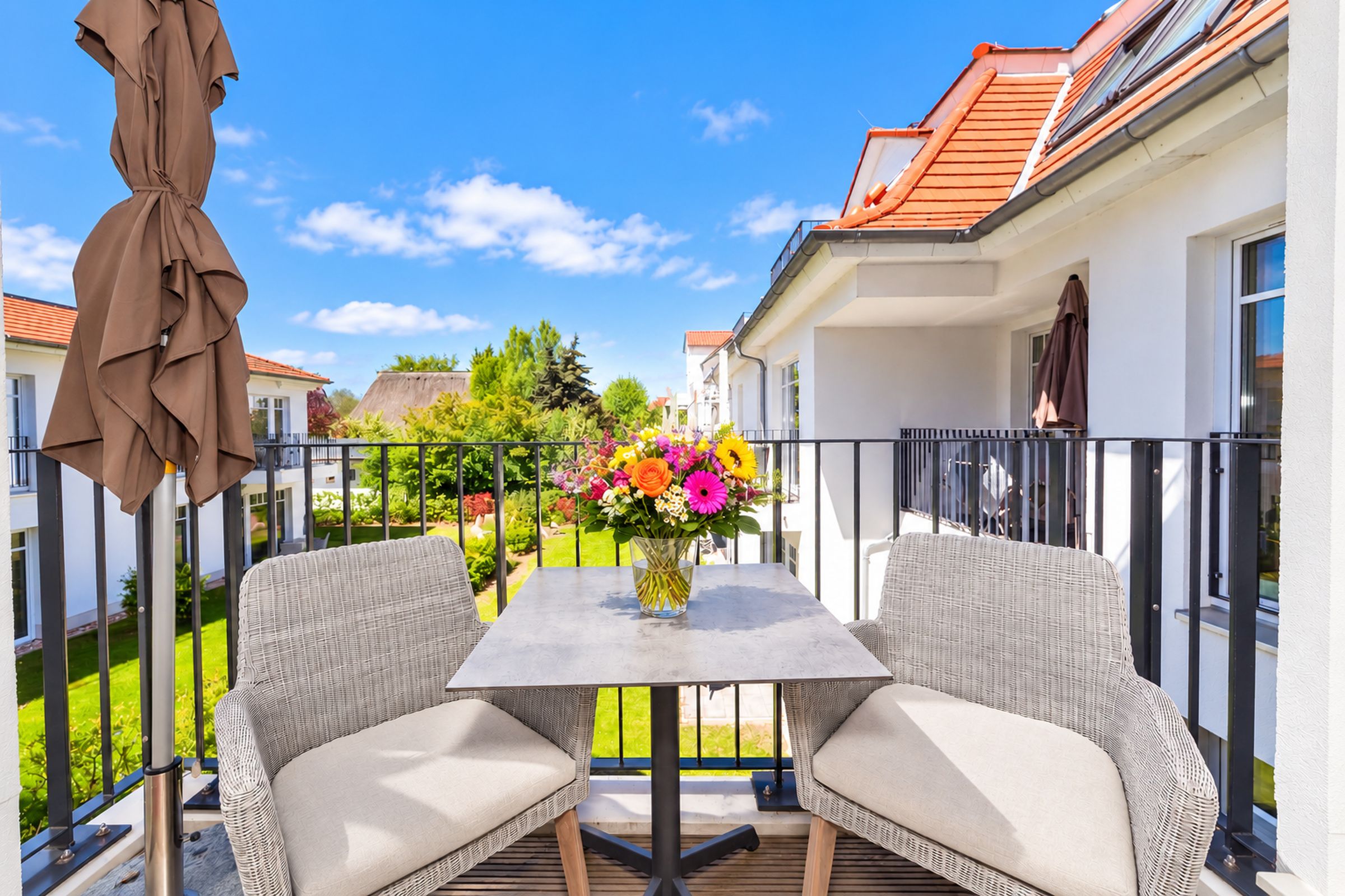 Un balcon avec deux fauteuils en osier, une table ornée d’un vase de fleurs et un parasol marron.