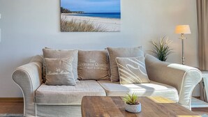 Un soggiorno con un divano color beige, un tavolino da caffè con una pianta e un quadro raffigurante una spiaggia.