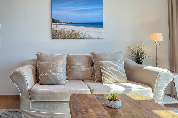 Ein Wohnzimmer mit einem beigen Sofa, einem Couchtisch mit Pflanze und einem Strandbild an der Wand.
