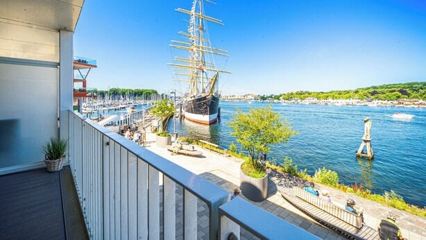 En balkong med utsikt över en hamn, ett stort segelfartyg och en staty.