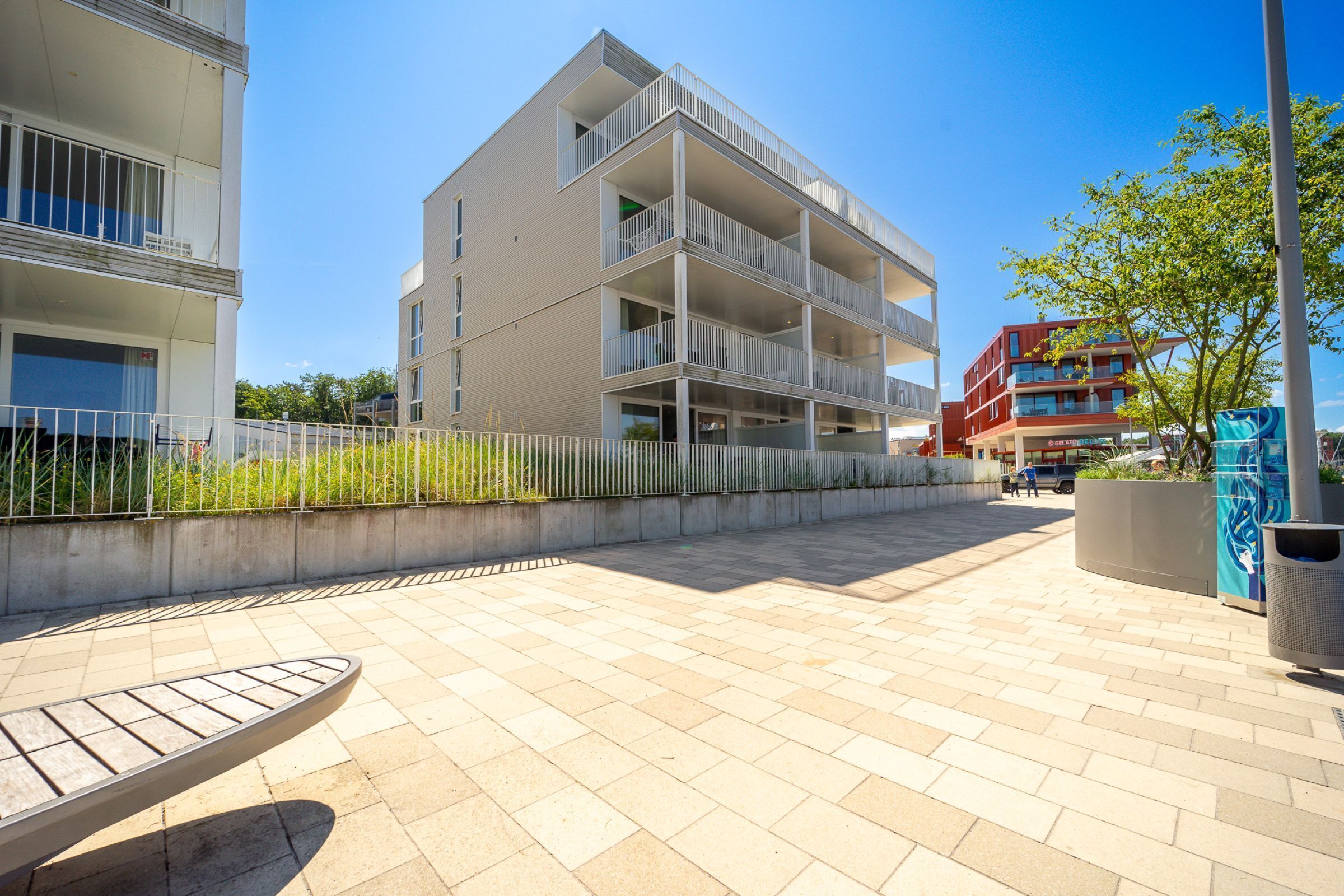 A modern apartment building with multiple balconies, a paved courtyard, and a nearby tree.
