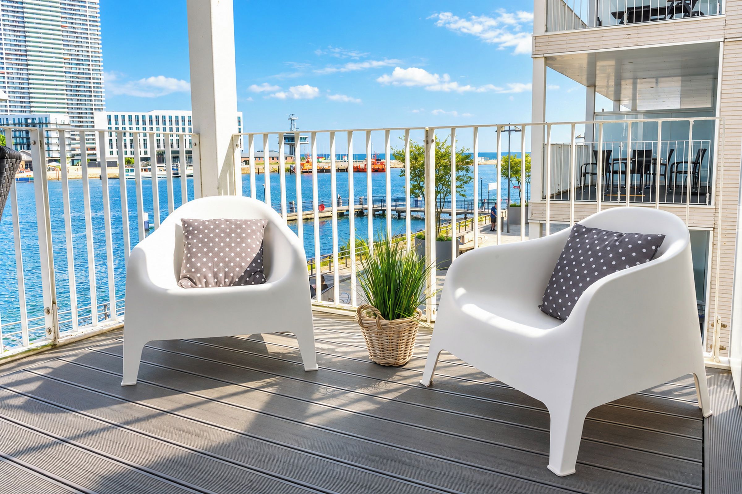 A balcony with two white chairs, one with a polka dot cushion and the other with a solid cushion, overlooking a marina with boats and a clear blue sky.