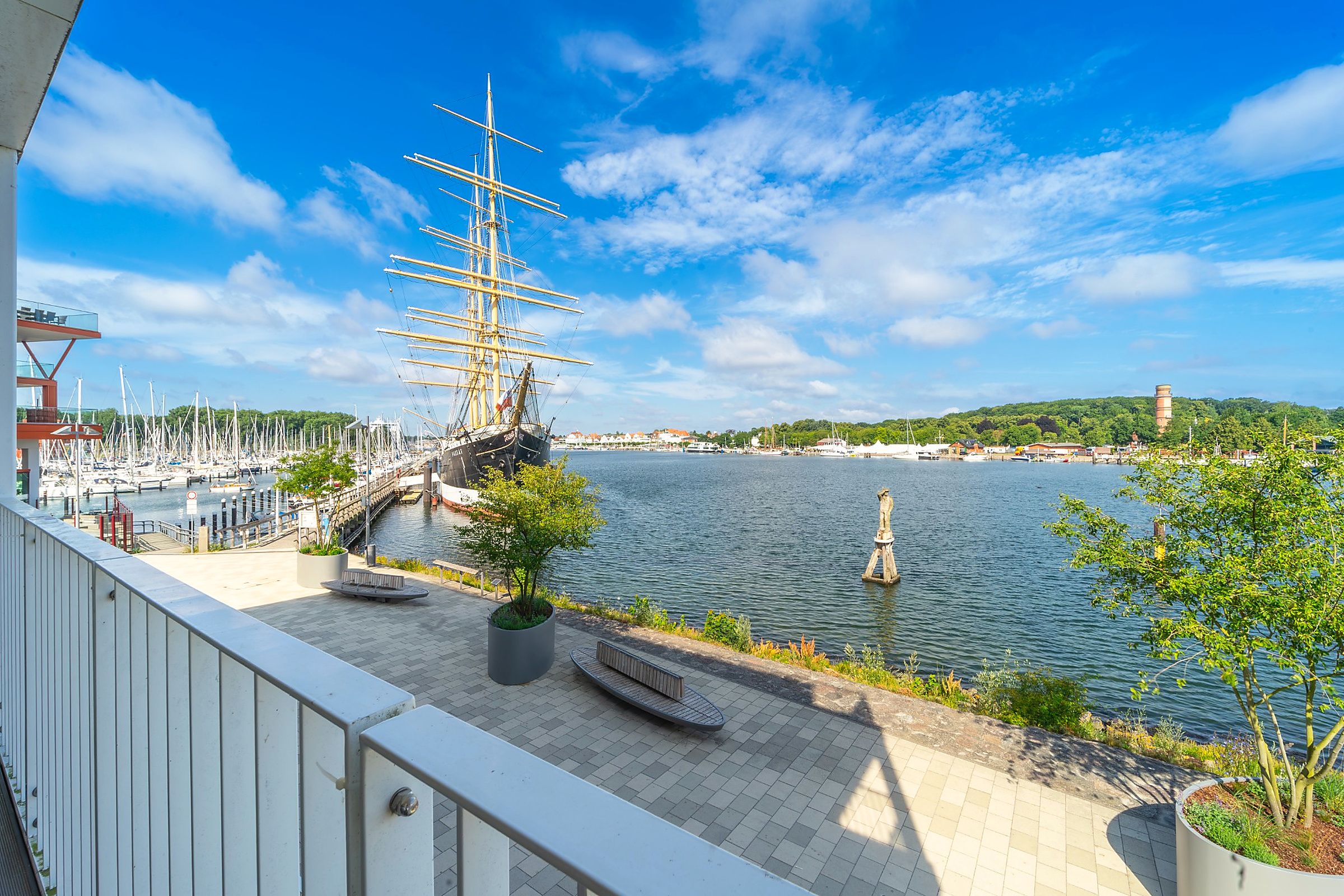 A waterfront promenade with a tall ship, potted plants, and a statue.