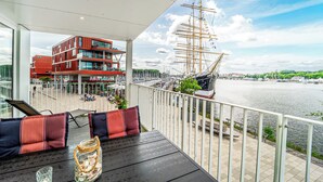 A balcony with a view of a river, a tall ship, and a modern building across the water. - Promenade Villa 03 Apartment 04 (Lübeck-Travemünde)