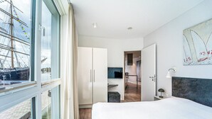 A modern hotel room with a large bed, a wardrobe, a desk with a lamp, and a view of a harbor with a ship. - Promenade Villa 03 Apartment 04 (Lübeck-Travemünde)