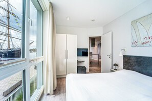 Een moderne hotelkamer met een groot bed, een garderobe, een bureau met lamp en uitzicht op een haven met een schip.