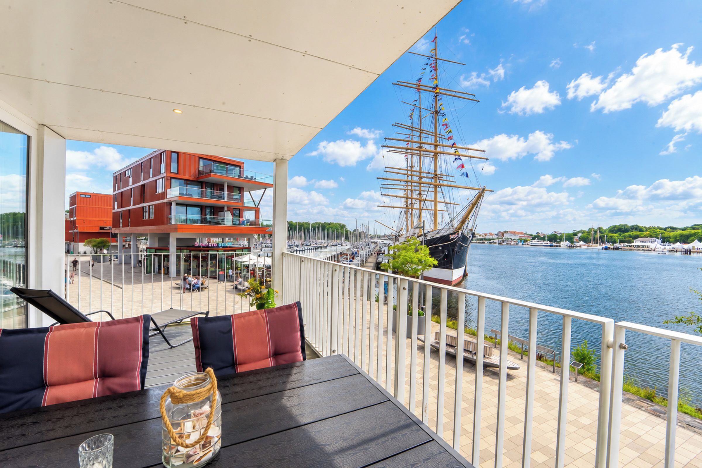 Un balcon offrant une vue sur un grand voilier et un bâtiment moderne situé de l’autre côté de l’eau.
