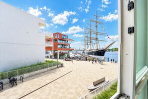 A waterfront promenade with a tall ship anchored nearby, a modern building with balconies, and a pedestrian walkway. - Promenade Villa 03, Apartment 04 (Lübeck-Travemünde)