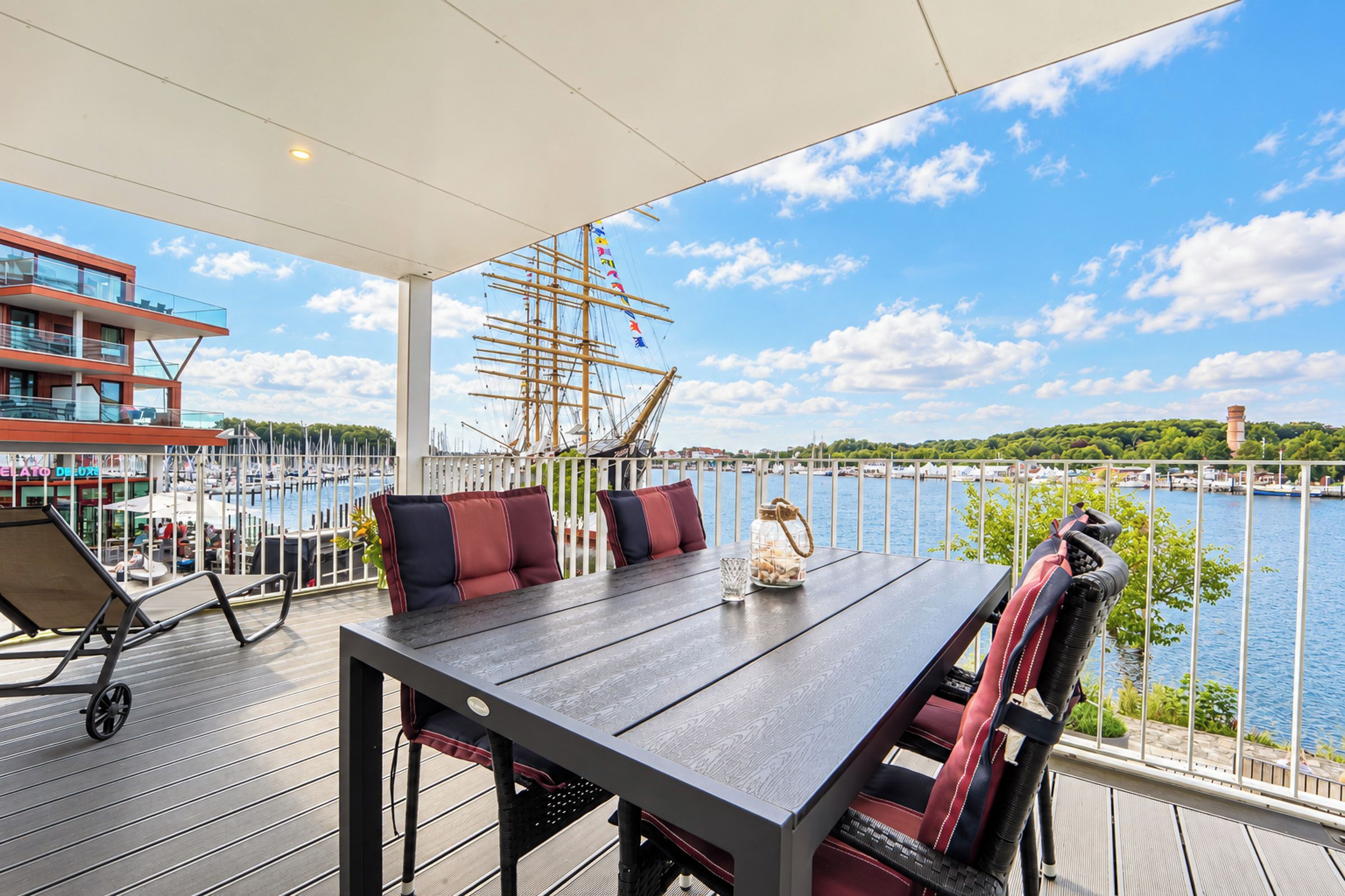 Um terraço com mesa e cadeiras, com vista para uma marina com barcos e um navio de grande porte.