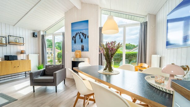 A modern dining area with a wooden table, white chairs, and a large window offering a view of greenery. - Vacation home Freibeuterweg 13 (Lübeck-Travemünde)