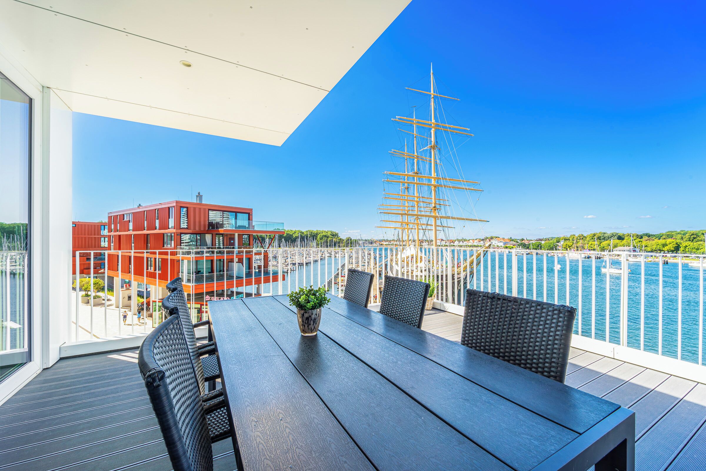 A balcony with a wooden table and chairs, a potted plant, and a view of a tall ship and water.