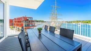 Een balkon met een houten tafel en stoelen, een bloempot en uitzicht op een hoge mast en water.