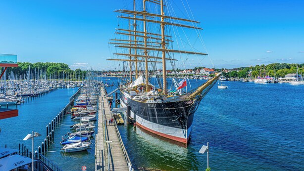 Een groot zeilschip aangemeerd in een jachthaven met diverse boten en een steiger.