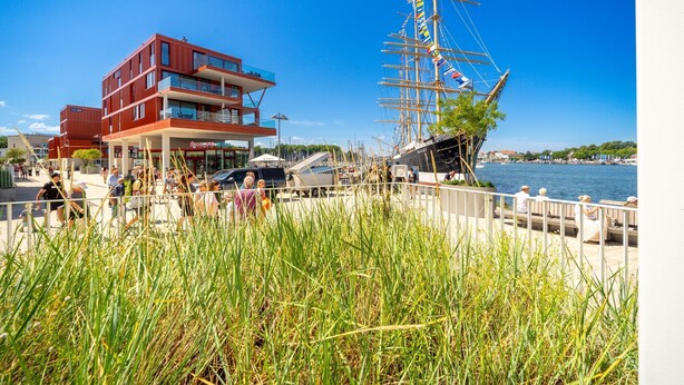 Ein Gebäude am Wasser mit modernem Design, ein großes Segelschiff liegt in der Nähe und im Vordergrund eine Rasenfläche.