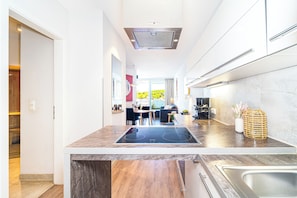 A modern kitchen with a central island, a built-in stove, and a large window offering a view of greenery outside. - Promenade Villa 03 Apartment 02 (Lübeck-Travemünde)