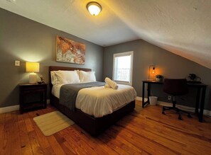 A bedroom with a bed, a desk, a chair, a lamp, a painting, and a window.