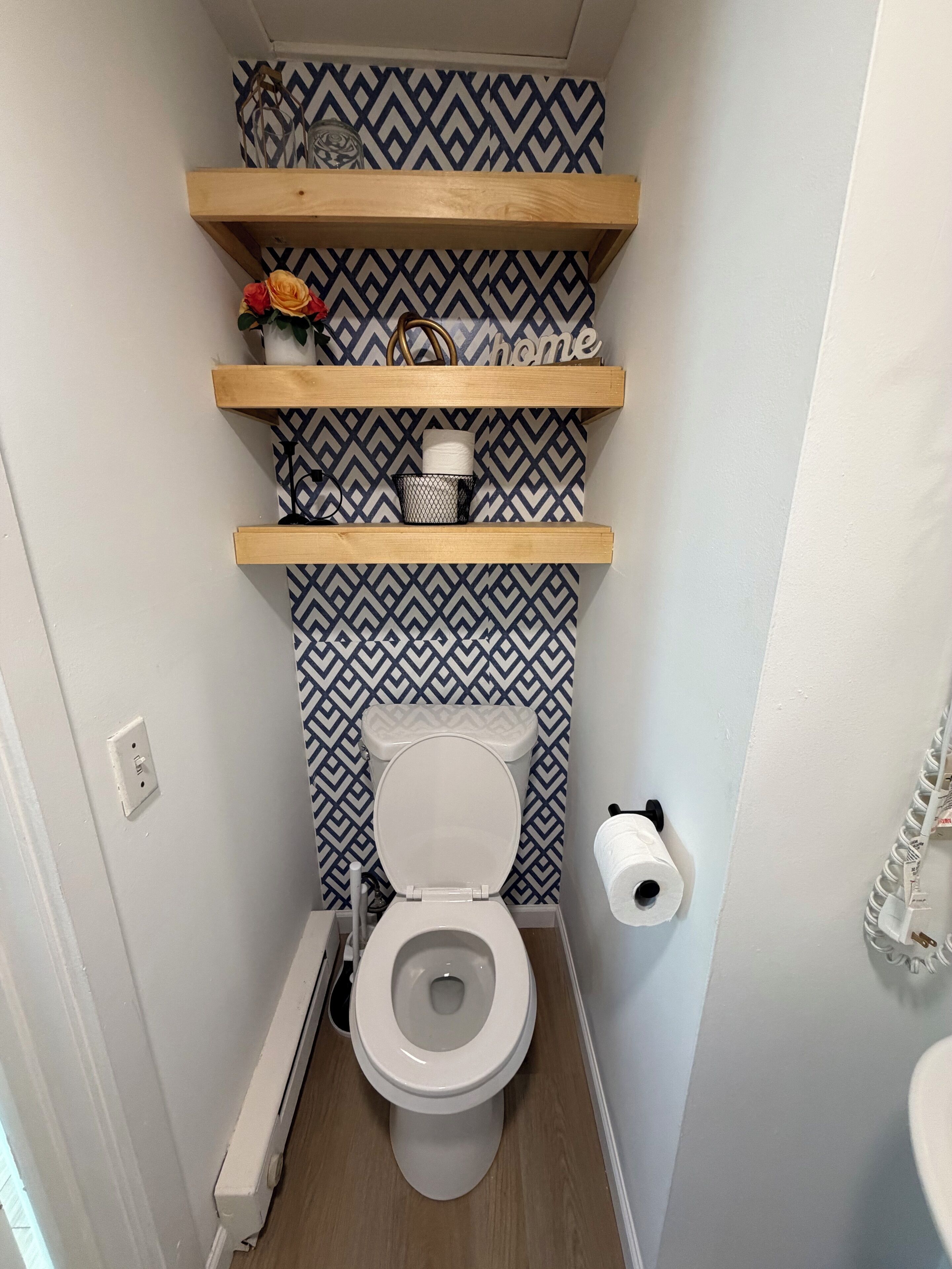 A small bathroom with a toilet, a shelf with toiletries, and a patterned wall.