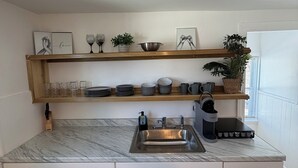 Una cuina moderna amb armaris blancs, tauleig de marbre, vàter d'acer inoxidable i cafetera. A la part superior del tauleig hi ha prestatges amb plats, gots i una planta.