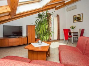 A cozy living room with a TV, wooden furniture, a large plant, and a dining area with red chairs. - Apartments Macolić Rab (Rab)