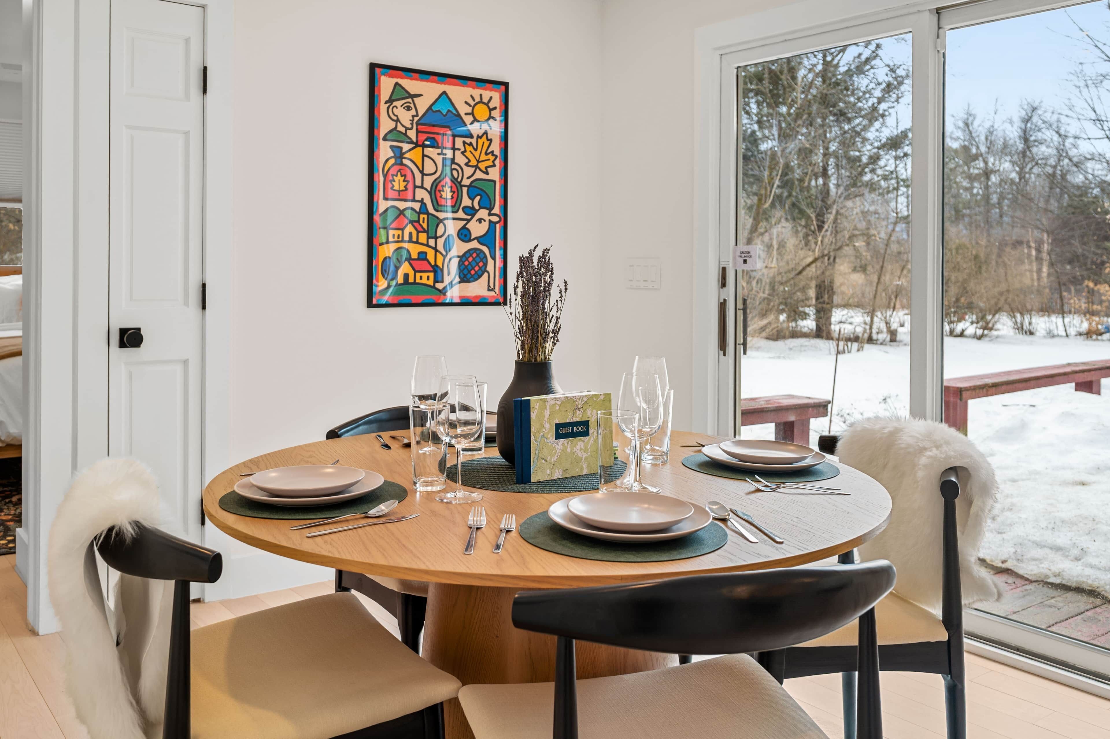 A dining area with a round table set for four, a colorful abstract painting on the wall, and a view of a snowy outdoor area through a glass door.
