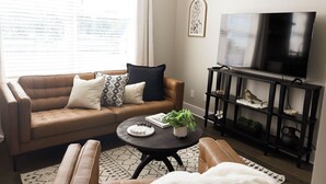 Un salon avec un canapé marron, une table basse ronde, un téléviseur posé sur un meuble noir et une étagère noire agrémentée d’objets décoratifs.