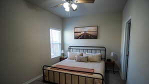 Un dormitorio con cama, ventilador de techo, ventana, mesita de noche y un cuadro en la pared.