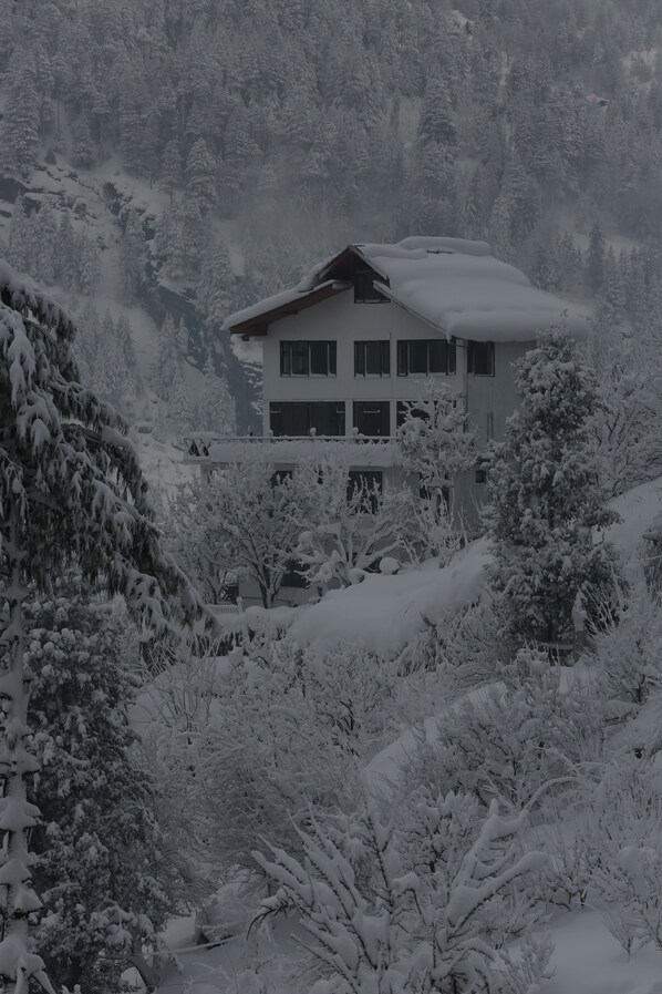 A two-story building with a snow-covered roof, surrounded by snow-laden trees and hills.