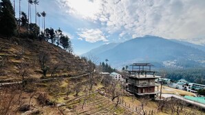 A mountainous landscape with a building featuring a green roof and a red flag on top. - North Star Manali (Manali)