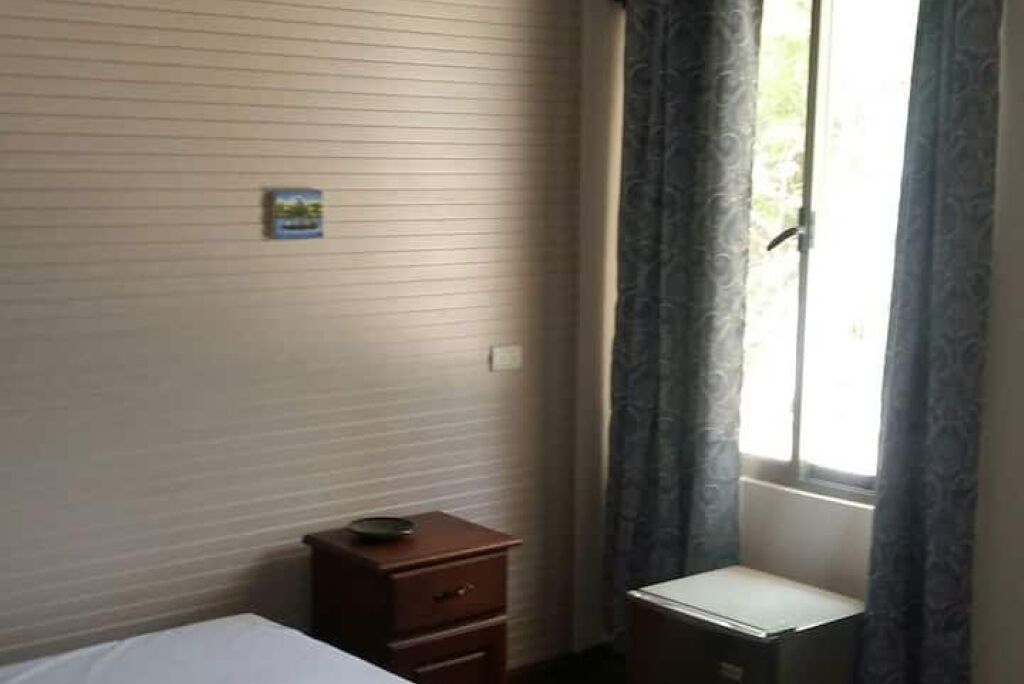 Een kleine, eenvoudige kamer met een bed, een houten nachtkastje, een klein kastje en een raam met gordijnen.