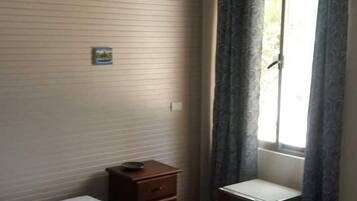 Een kleine, eenvoudige kamer met een bed, een houten nachtkastje, een klein kastje en een raam met gordijnen.