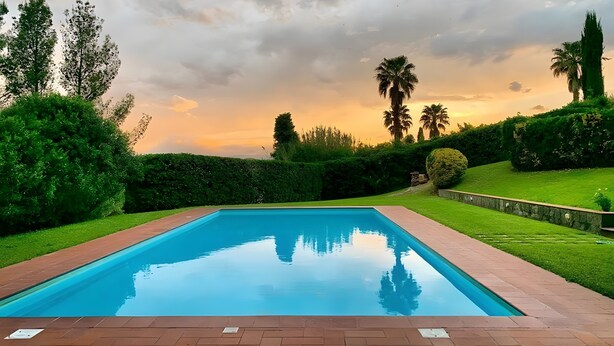 Una piscina rectangular con borde de ladrillo, rodeada de setos y árboles bien cuidados, bajo un cielo nublado.