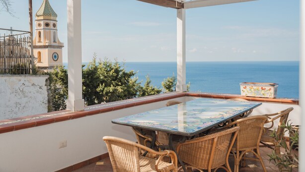 Un espace extérieur abrité, doté d’une table et de chaises, offrant une vue sur la mer et, au loin, une tour.