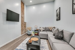A modern living room with a grey sofa, a flat-screen TV mounted on the wall, a coffee table with a woven coaster, and a patterned rug. - Gateway to Mammoth Cave! Hike, Bike & Explore (Horse Cave)