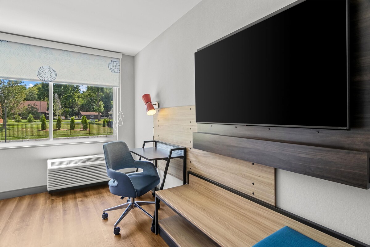 A modern office space with a large flat-screen TV mounted on the wall, a desk with a chair, and a view of a green outdoor area through a large window.