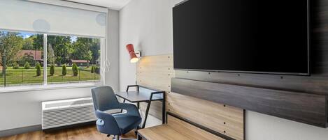 A modern office space with a large flat-screen TV mounted on the wall, a desk with a chair, and a view of a green outdoor area through a large window.