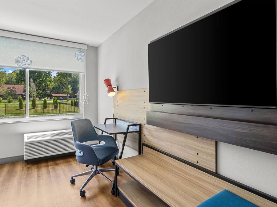 A modern office space with a large flat-screen TV mounted on the wall, a desk with a chair, and a view of a green outdoor area through a large window.