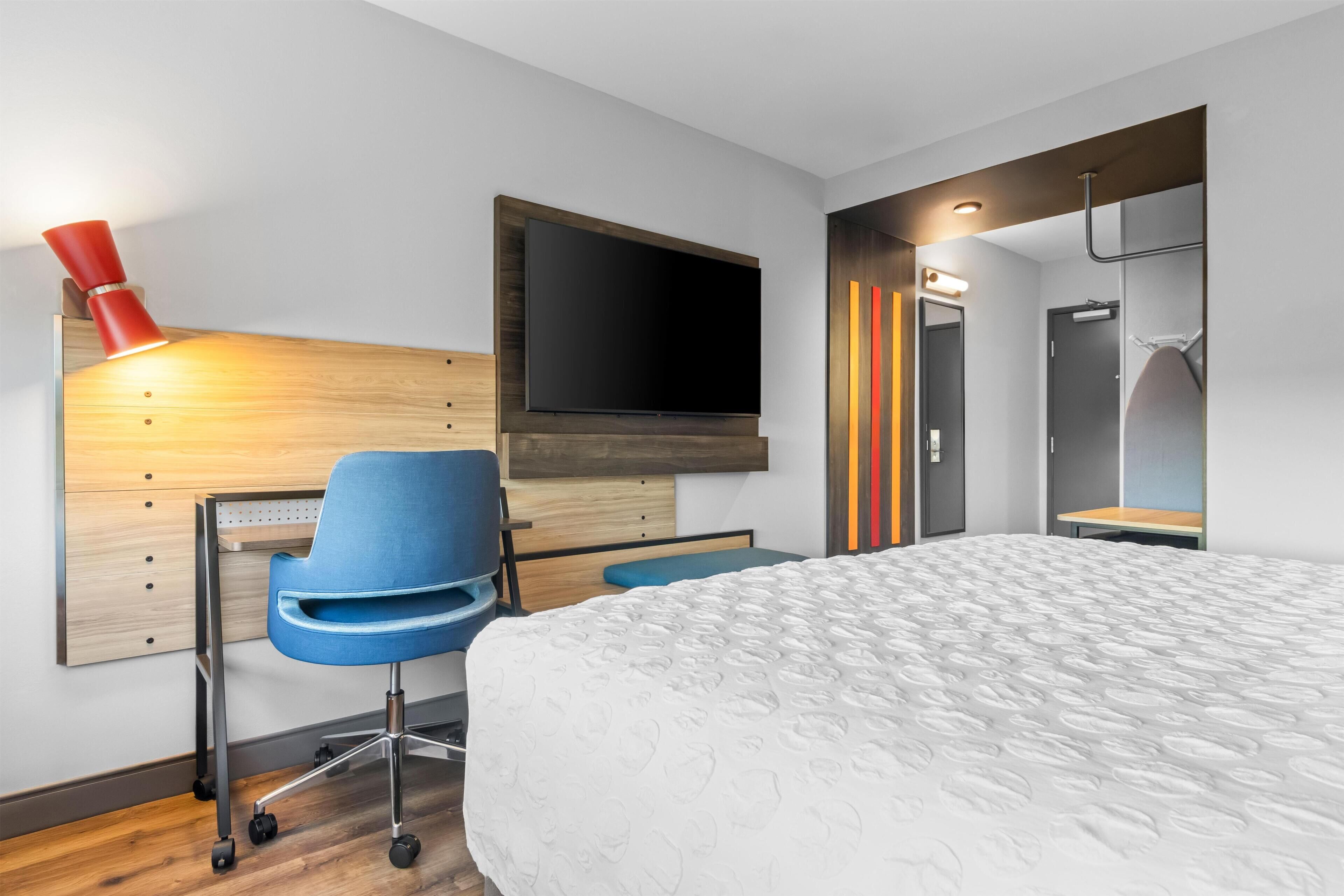 Una habitación de hotel moderna con una cama, un escritorio con silla, un televisor montado en la pared y un baño con espejo y toallero.